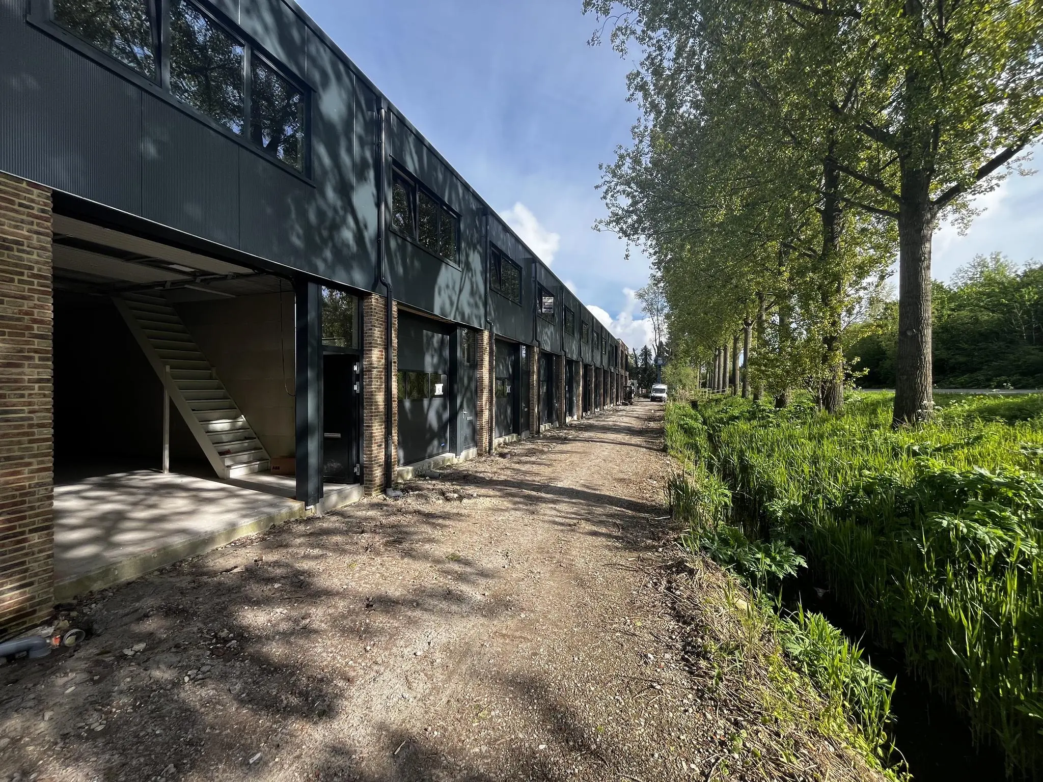 Nieuw bedrijfsgebouw aan de Broekdijk West met open units, gelegen langs een onverhard pad en een groen watertje met hoge bomen.